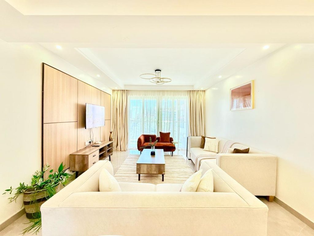 Living area furnished with beautiful couches and a mounted smart tv and a Tv Console...there is also a coffee table and large windows covered by sheer white curtains at Fully Furnished 3 bedroom Apartment in Westlands