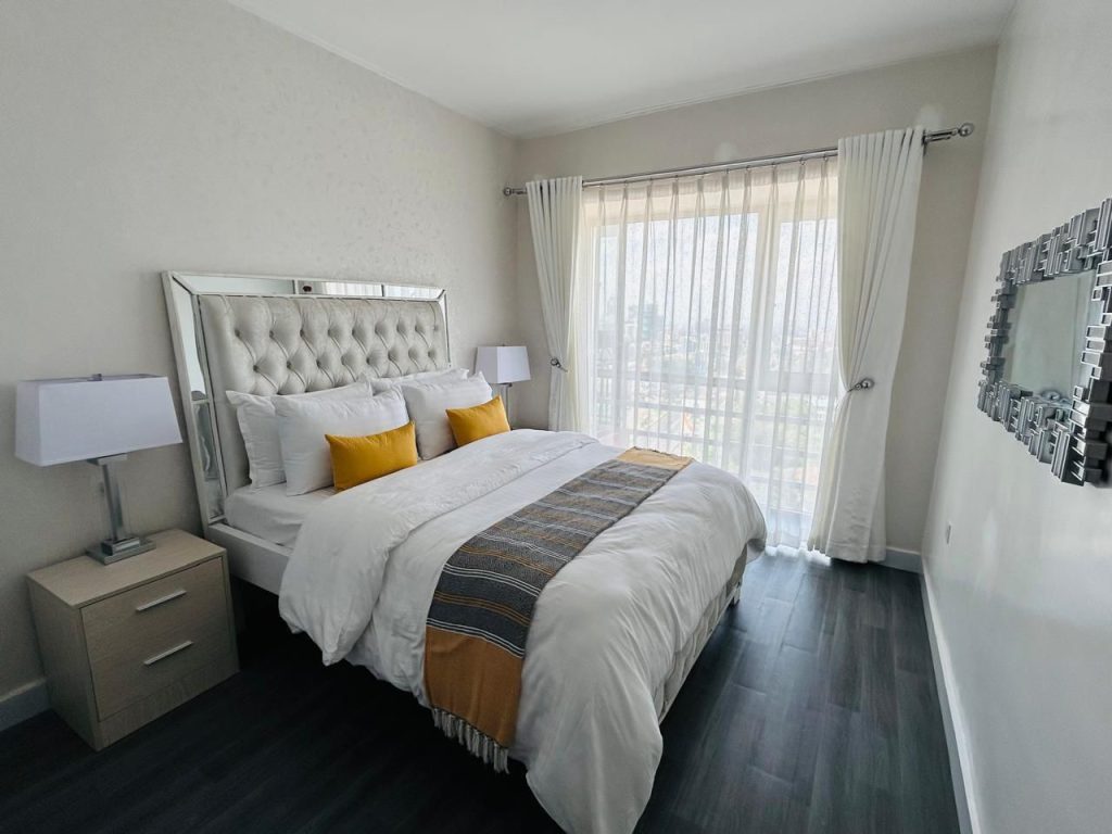 king size bed with white linen and yellow throw pillows and the view of the large windows with curtains at Kilimani 2 bedroom Airbnb with swimming pool