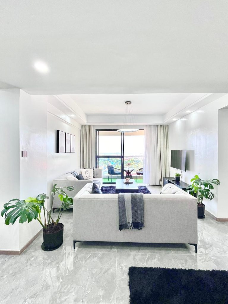 the living area of the Two Bedroom Furnished Apartment for rent in Nairobi with couches, a mounted smart Tv, a coffee table and the view of the balcony through the large windows