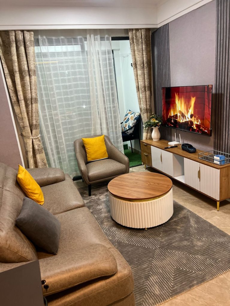 The living room of the Furnished Apartment with Rooftop Terrace in Kilimani with brown couches, a coffee table, a mounted smart tv and and large windows with curtains