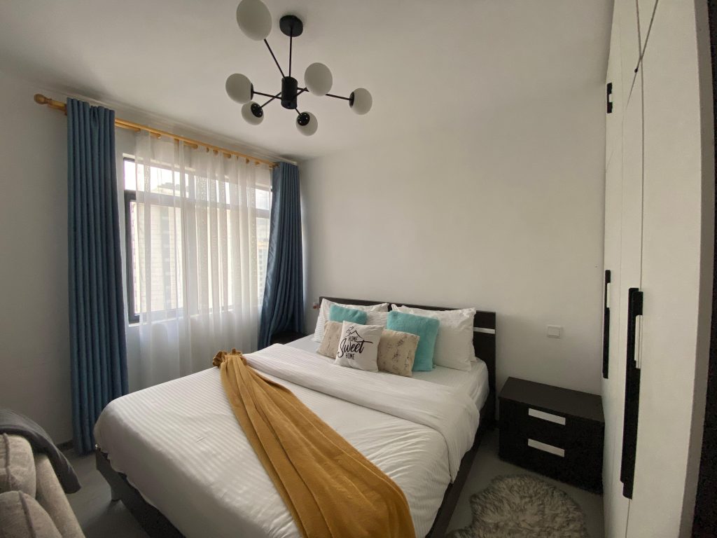 A queen size bed with white covers and large windows with curtains at Furnished Studio Apartment in Kilimani