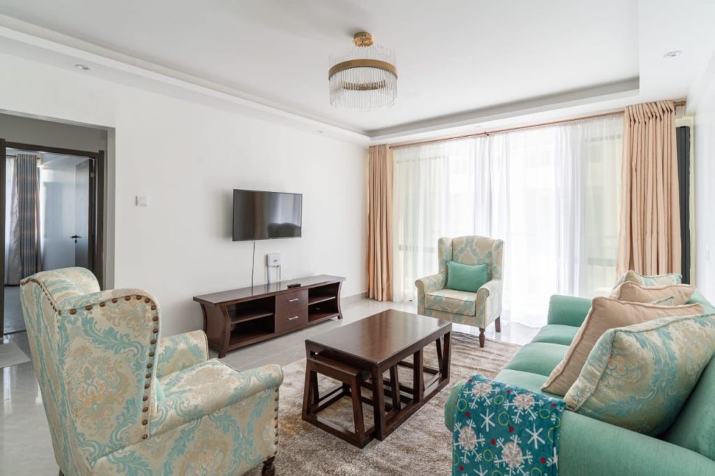 a beautiful living room with couches, a coffee table ,a mounted smart Tv with a console and large windows covered in transparent white sheers at Extended Stay Furnished Apartment in Nairobi