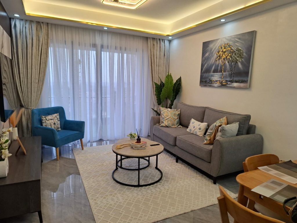 Beautiful living area in 2 Bedroom Corporate Housing in Kilimani Nairobi featuring a grey 2-seater couch, blue one-seater couch, and large windows with natural light.