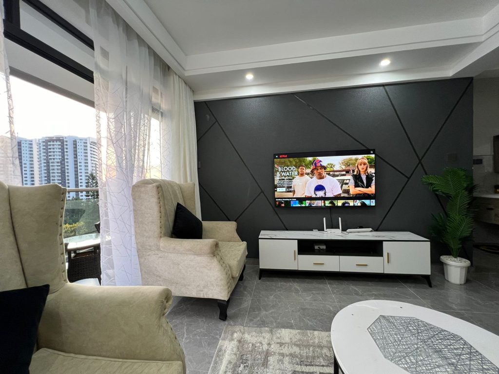 Spacious sitting area in 1 Bedroom Furnished Apartment in Kileleshwa featuring a one-seater couch, smart TV, and large windows opening to a balcony.