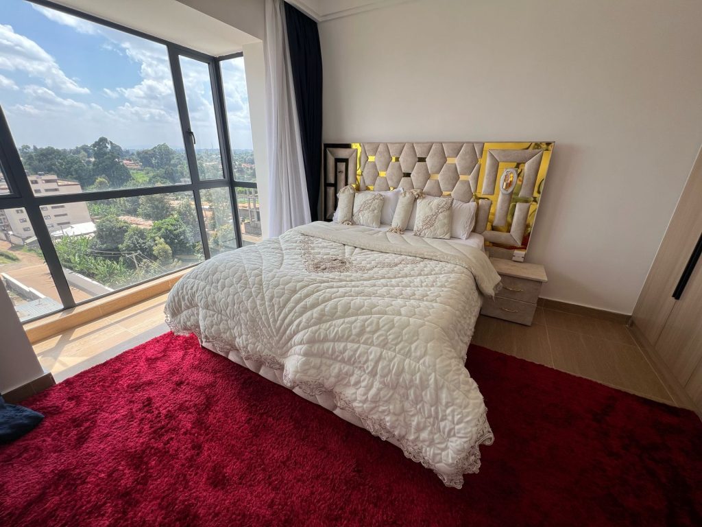 Spacious master bedroom in Ruaka furnished daily and monthly rental in Nairobi with a queen size bed, red carpet, and natural lighting from large windows.