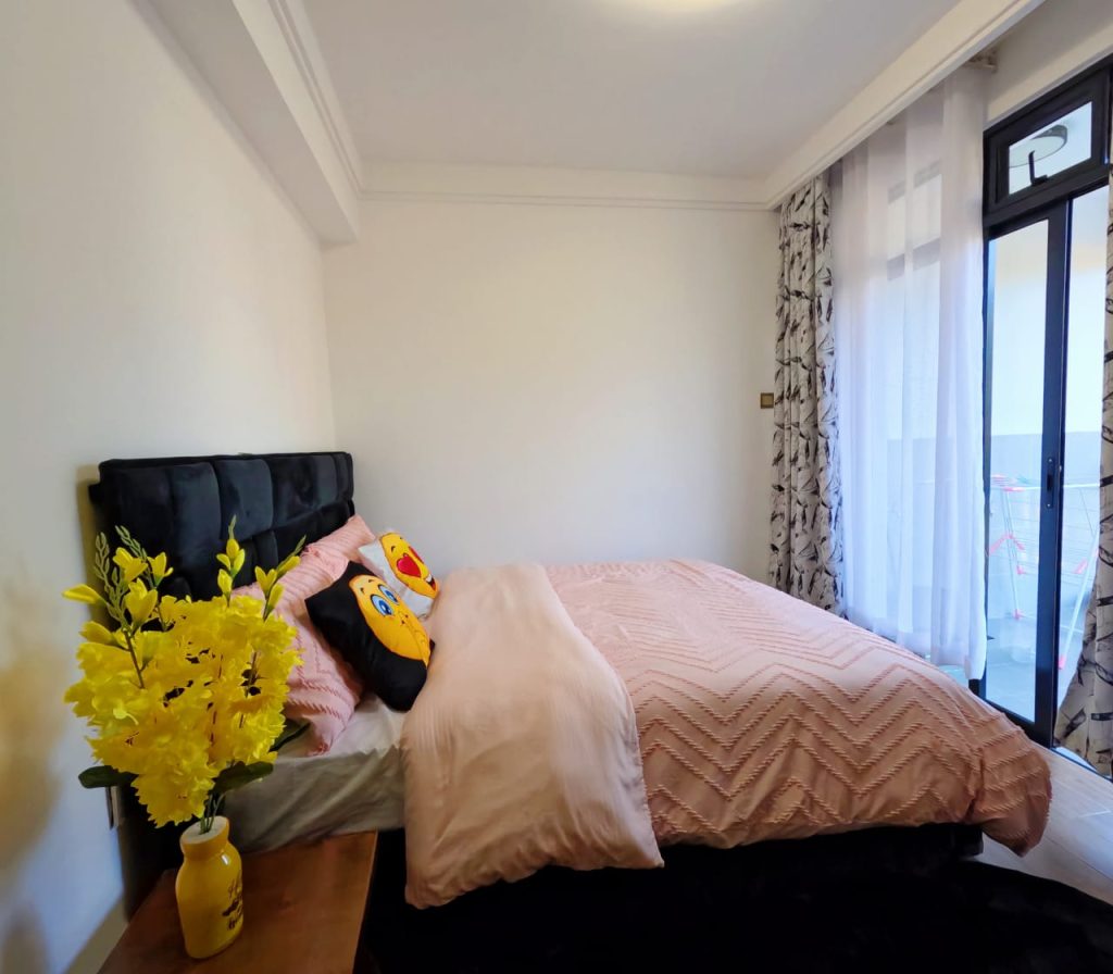 Queen size bed with pink duvet next to large windows opening to a balcony in a serviced furnished 1‑bedroom apartment in Lavington.