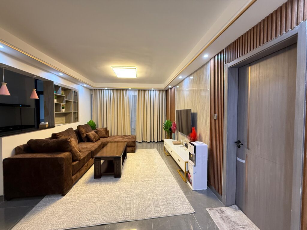 warm lounge of the Furnished Apartment at Alina Harbour Kilimani with a brown sofa set a coffee table, a mounted smart Tv and an artistic shelving.